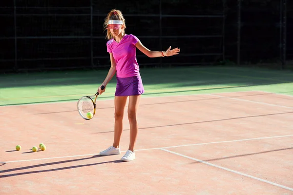屋外テニスコートでのトレーニング中にキャップとテニスユニフォームの若い女性がテニスボールを提供しています。スポーツウェアのきれいなスポーティな女の子は日没時にテニスの訓練を持っています. — ストック写真