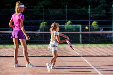 Beyaz üniformalı küçük kız koç ile tenis eğitimi sırasında net altında topa isabet. Çekici kadın antrenör gün batımında açık sahada küçük tenisçi ile egzersizler yapıyor.