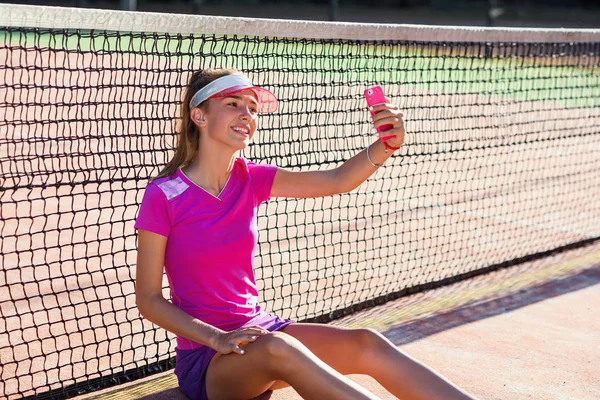 ピンクの制服を着た若い美少女とスポーティなキャップがネット近くのテニスコートに座り、日没時にスマートフォンで自撮り写真を撮る。テニスの練習後やスマートフォンの使用後のリラックス. — ストック写真