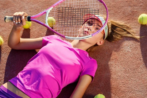 Top view of cute girl in the cap lying on tennis outdoor court with racket in the hand and a lot of balls on the ground after training.