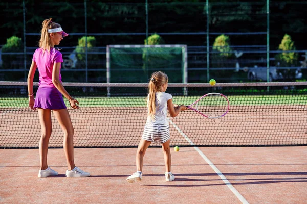 コーチとのテニストレーニング中にネットの下に白いユニフォームの小さな女の子。魅力的な女性コーチは、日没時に屋外コートで少しテニスプレーヤーとエクササイズを行います. — ストック写真