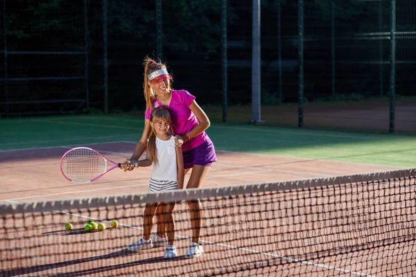 Liten flicka i vit Uniform slå bollen undernätet under tennis träning med tränare. Attraktiv kvinnlig tränare gör övningar med liten tennisspelare på utomhusterrassen vid solnedgången. — Stockfoto