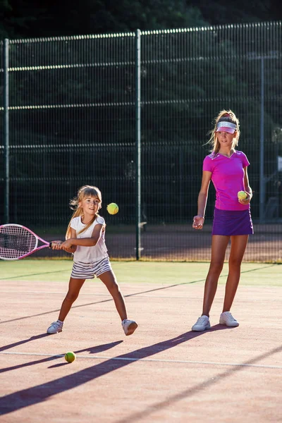 Liten flicka i vit Uniform slå bollen undernätet under tennis träning med tränare. Attraktiv kvinnlig tränare gör övningar med liten tennisspelare på utomhusterrassen vid solnedgången. — Stockfoto