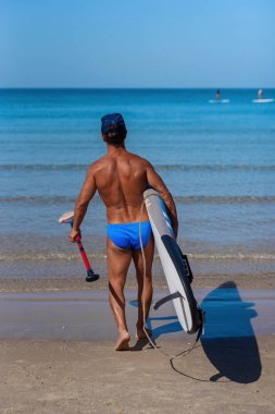 Elinde sörf tahtası ve kürek ile deniz sahilinde tabaklanmış adam suya girer. Kavram sportif ve sağlıklı bir yaşam tarzıdır.