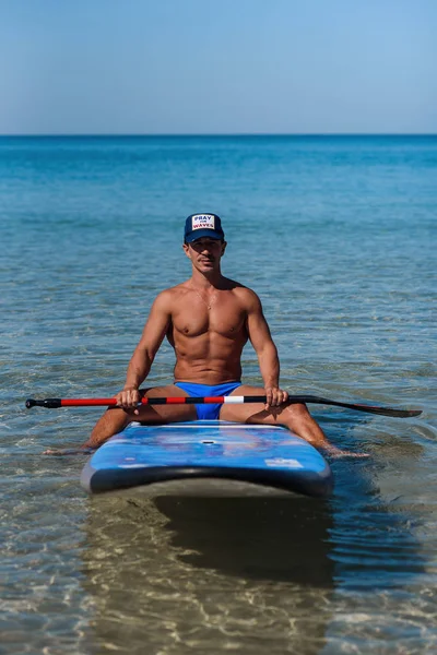 O homem bronzeado senta-se na praia do mar perto de sua prancha de surf e olha para a câmera. O conceito é um estilo de vida desportivo e saudável . — Fotografia de Stock