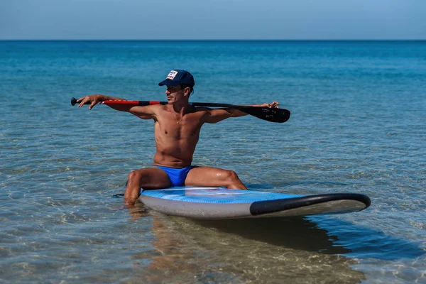 Tanned sporty man in a cap sits on his surfboard on the water holding by hands an oar behind his head and looks into the water. The concept is a sporty and healthy lifestyle.