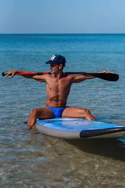 Tanned sporty man in a cap sits on his surfboard on the water holding by hands an oar behind his head and looks into the water. The concept is a sporty and healthy lifestyle.