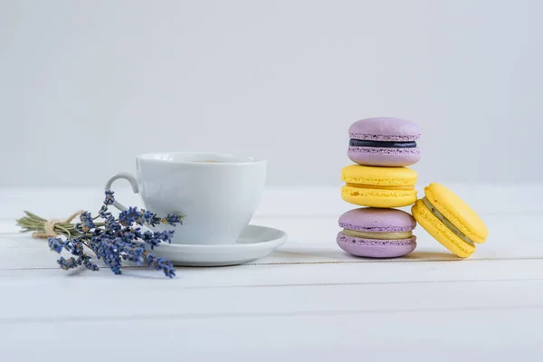 Delicious violet and yellow macarons and cup of latte or americano and branch of fragrant lavender on white wooden background. — Stock Photo, Image