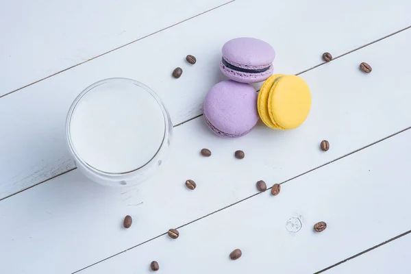 Sweet delicious violet and yellow macarons and cup of latte or americano and scattered coffee beans on white wooden background. — Stock Photo, Image