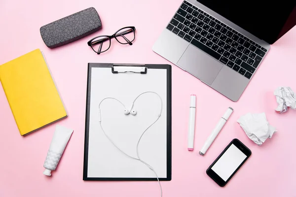 Womans pink office desk with headphones laid out like a heart. Womans workplace with laptop, smart phone with blank white screen, a cream, lipstick, eyeglasses and supplies.