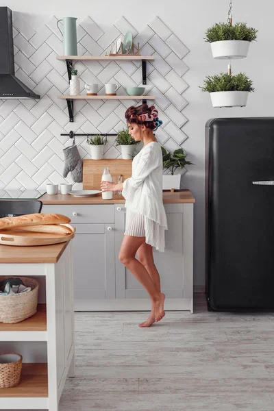 Elinde bir şişe süt tutan sevimli güzel bir kadın. Güzel kız modern rahat mutfakta kahvaltı hazırlanıyor.