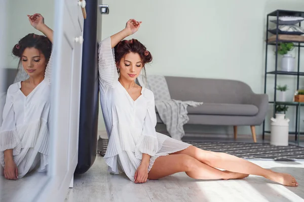 Jovem mulher sentada no chão em casa na cozinha. Uma mulher adorável com cabelo ondulação vestida de roupão branco tendo descanso pela manhã . — Fotografia de Stock