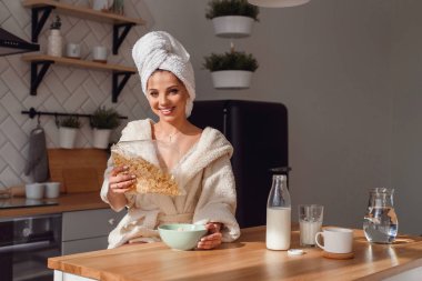 Güzel genç kadın süt ile tabakta mısır gevreği döker. Kız sabah şık rahat mutfakta sağlıklı bir kahvaltı var.