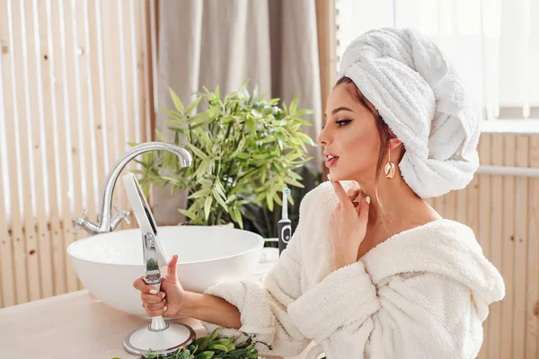 Hermosa joven con una toalla blanca en la cabeza vestida con albornoz se mira en el espejo en el baño . — Foto de Stock