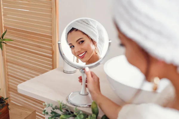 Mujer joven atractiva con una toalla blanca en la cabeza vestida con albornoz se mira en el espejo en el elegante baño después de la ducha de la mañana . — Foto de Stock