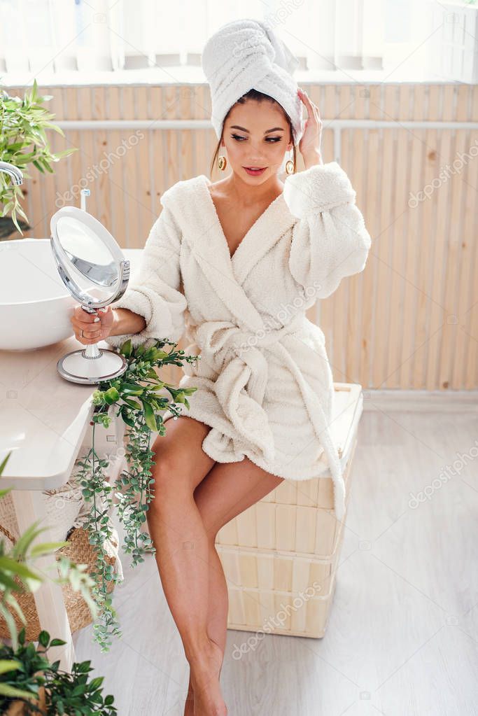 Pretty young girl with a white towel on head dressed in bathrobe looks at herself in the mirror in cozy bathroom after morning shower.