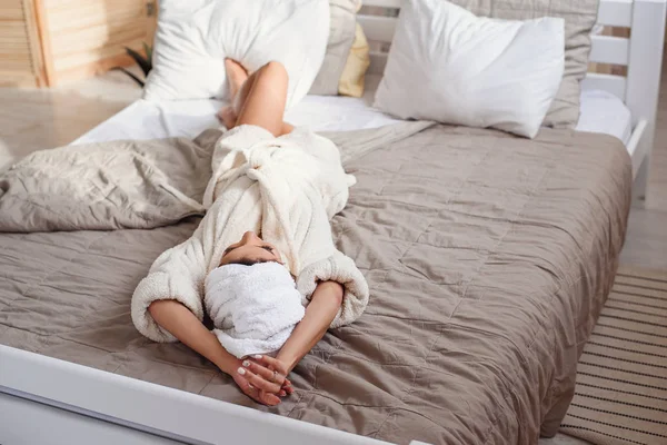 Bela jovem deita-se na cama confortável em casa após o procedimento de spa manhã vestida com roupão de banho branco. Menina bonita ter relaxar na cama de manhã . — Fotografia de Stock