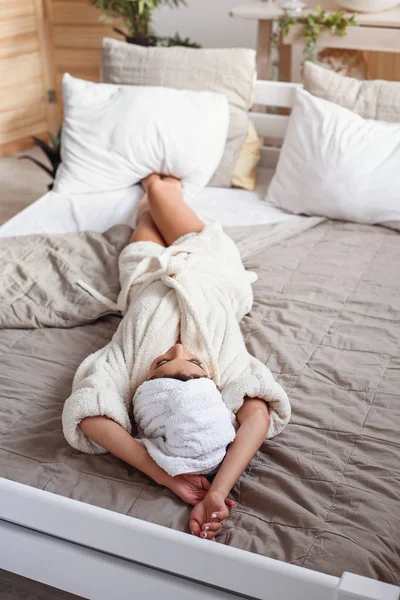 白いバスローブを着たお風呂の後、自宅のベッドに横たわっている魅力的な笑顔の若い女性。朝ベッドでくつろぐ美少女. — ストック写真
