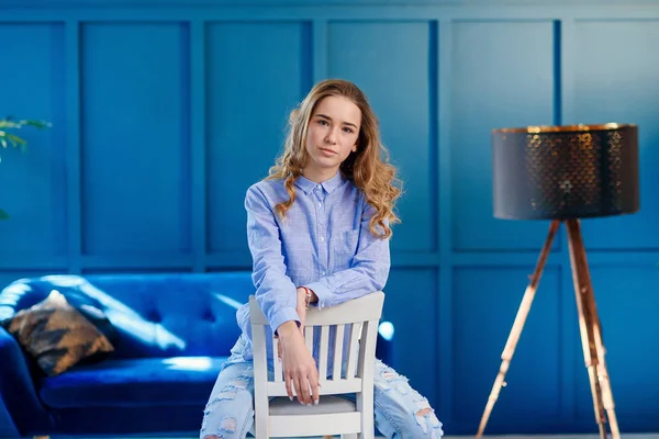 Menina elegante bonita em roupas casuais senta-se em uma cadeira de madeira branca no interior moderno elegante com fundo azul . — Fotografia de Stock