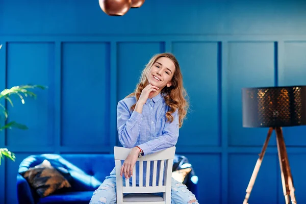 Menina elegante bonita em roupas casuais senta-se em uma cadeira de madeira branca no interior moderno elegante com fundo azul . — Fotografia de Stock