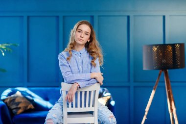 Rahat giysiler içinde güzel şık kız mavi arka plan ile şık modern iç beyaz ahşap sandalyede oturur.