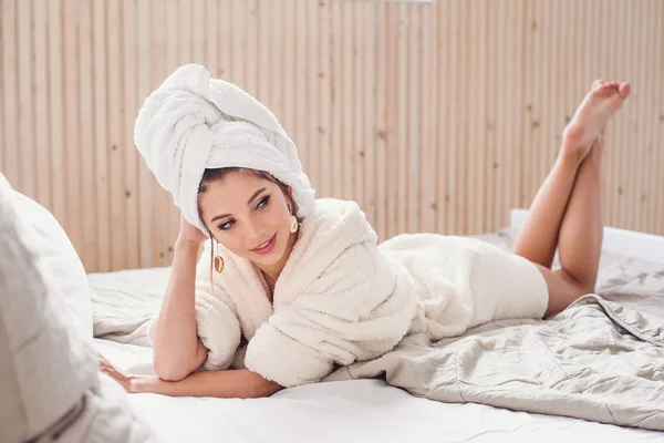 Förförisk leende ung kvinna som ligger på sängen hemma efter morgonen Spa rpocedures klädd i vit badrock. Vacker flicka som har slappna av på sovrummet på morgonen. — Stockfoto