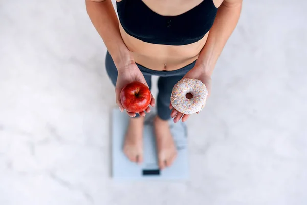 Sporty woman with a perfect body measuring body weight on electronic scales and holding a donut and a red apple. Unhealthy food, healthy low calorie meals. Diet, a healthy lifestyle.