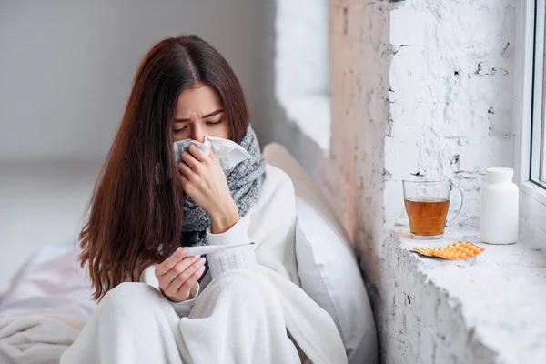Sick woman caught cold, feeling illness and sneezing in paper wipe. Closeup of beautiful unhealthy girl covered in blanket wiping nose and looking at thermometer. Healthcare concept.