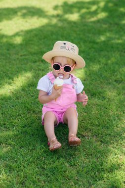 Mutlu bebek kız giyinmiş pembe yaz giysileri, sarı şapka ve pembe güneş gözlüğü yeşil çim üzerinde oturur ve güneşli bir bahçede beyaz dondurma yiyor.