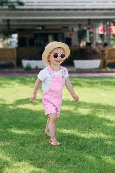 Stylish girl in a pink overalls, hat and pink glasses walks on the green grass. Summer vacation concept. — Stock Photo, Image