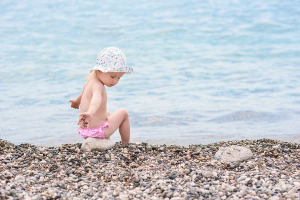 Cute toddler girl sits on the sea beach in hot summer day and plays with stones.