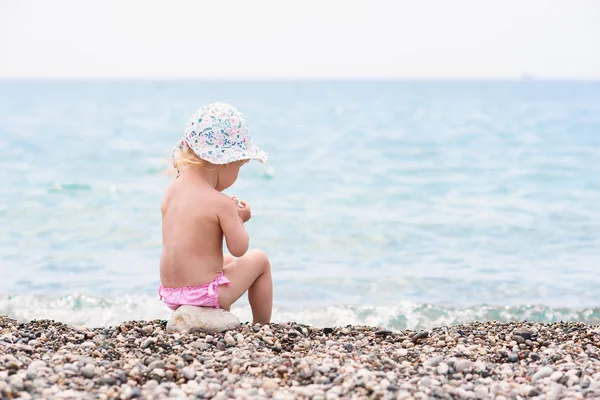 Carino bambino ragazza si siede sulla spiaggia del mare in calda giornata estiva e gioca con le pietre . — Foto Stock