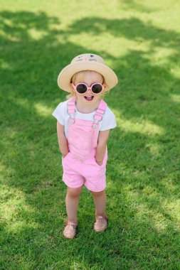 Şirin elbise, şapka ve pembe Gözlük Sevimli kız güneşli yaz gününde yeşil çim ile çim üzerinde duruyor.