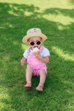 Mutlu bebek kız giyinmiş pembe yaz giysileri, sarı şapka ve pembe güneş gözlüğü yeşil çim üzerinde oturur ve güneşli bir bahçede beyaz dondurma yiyor.
