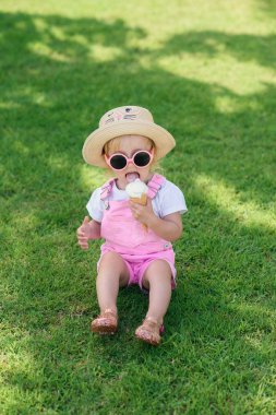 Pembe yaz tulum giyen komik mutlu yürümeye başlayan kız, şapka ve pembe güneş gözlüğü güneşli bir bahçede vanilya beyaz dondurma yiyor yeşil çim oturur.