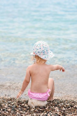 Deniz plajında otururken yaz şapka mutlu yürümeye başlayan kız su içine taş atar.
