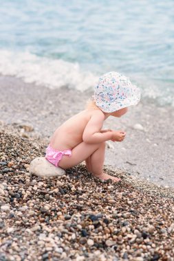 Sevimli Toddler kız sıcak yaz gününde deniz plajında oturur ve taşlar ile oynuyor.