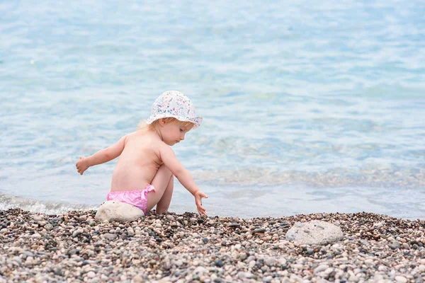 Cute toddler girl sits on the sea beach in hot summer day and plays with stones.
