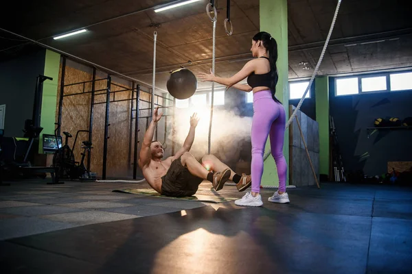 Caucásico hombre deportivo haciendo ejercicio de pelota abdominal con entrenador femenino personal en el gimnasio . — Foto de Stock