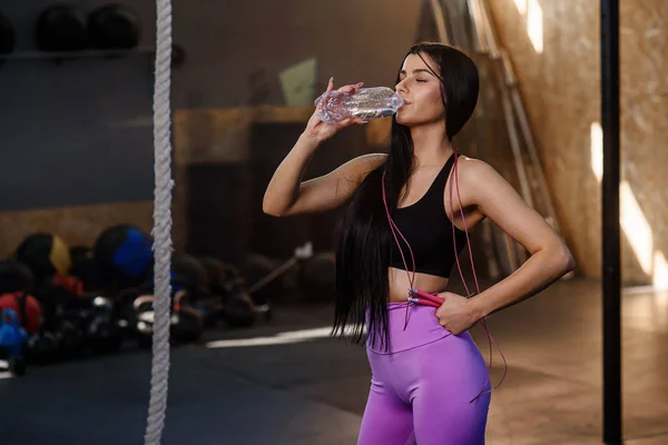 Beautiful fit woman drinking water from plastic bottle after workout in the gym.