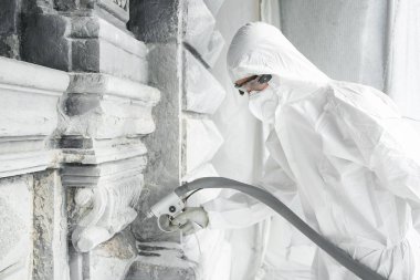 Beyaz koruyucu üniformalı adam, taş tan oyulmuş heykeli topraktan ve betondan kumlama makinesiyle temizler. Taş heykel restorasyonu. Yüksek basınç altında bir kum jeti.