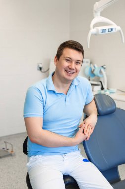 Dişçi sandalyede oturuyor, poz arıyor, kameraya bakıyor. Modern ekipman ile özel diş hekimliği kliniğinde çalışan mavi ve beyaz üniforma giyen sakallı adam.