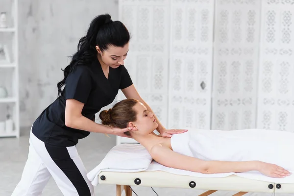 Jovem massagista profissional fazendo massagem para mulher caucasiana sonolenta no fundo da sala branca . — Fotografia de Stock