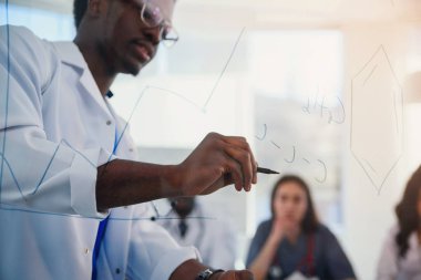 Genç erkek Afrikalı doktor cam tahtaya moleküler formül çiziyor. Laboratuarında öğrencisi için şeffaf bir tahtaya kimyasal formüller yazan bir öğretmen..