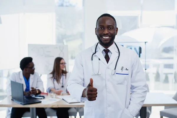 Portrait of smiling afro american male doctor showing thumb up sign. Medical assistant or student in white uniform with stethoscope. Handsome male doctor with a medical group at meeting room.