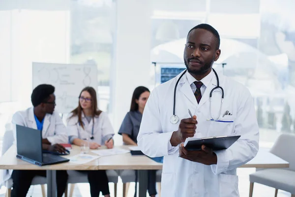 Handsome African doctor in white coat makes some notes. Young medical student with a stethoscope around his neck holds a folder.