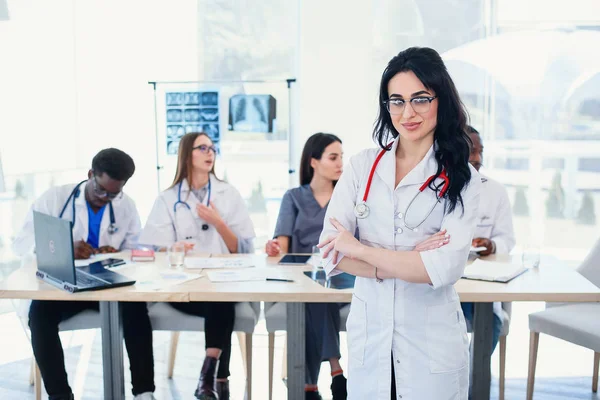 Smiling medical doctor woman with stethoscope standing in front of medic team at hospital. Attractive young female doctor in white coat and glasses looking at the camera.