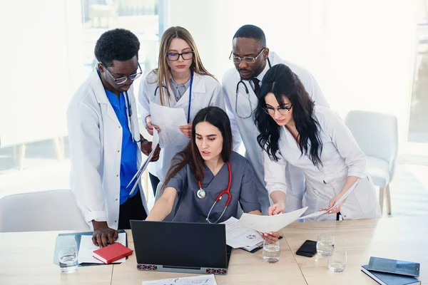 Multinational group of doctors looks at something in the laptop. A female doctor is sitting at the desk. Doctors work together in one hospital.