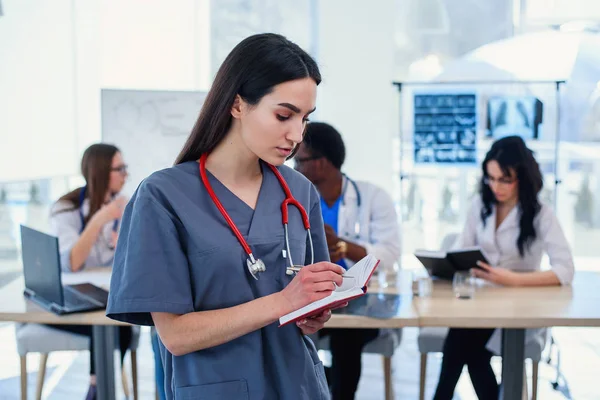 Female doctor in grey uniform with stethoscope on the neck in medical clothes smiling to the camera and crossing hands while medical team working on the background in modern hospital.