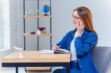 Denim gömlek giymiş kadın tatil gezisi planlama sırasında masada oturur akıllı telefon kullanır.